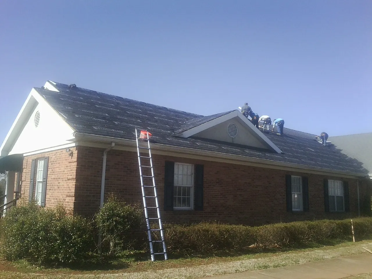 Crew tearing shingles off a brick ranch roof with a ladder set — Emergency Roof Repair work by our crew in Point Clear