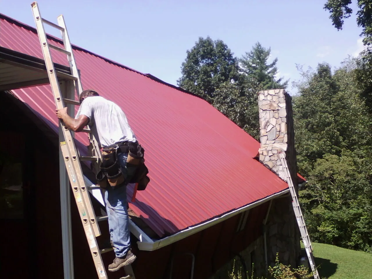 Roofer on a ladder beside a red metal roof and stone chimney — Soffit & Fascia Repair by Roofing Brothers in Point Clear